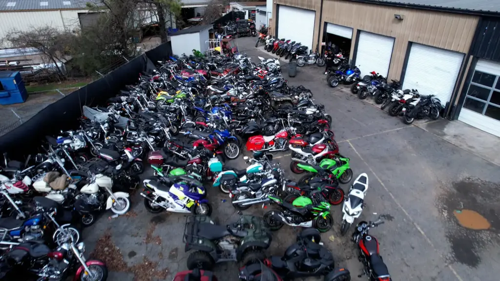 Dozens of motorcycles are parked close together in a parking lot outside a large building with open garages.