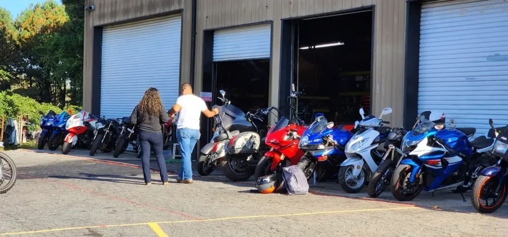 Two people stand beside sport motorcycles parked outside a garage with partially open doors.