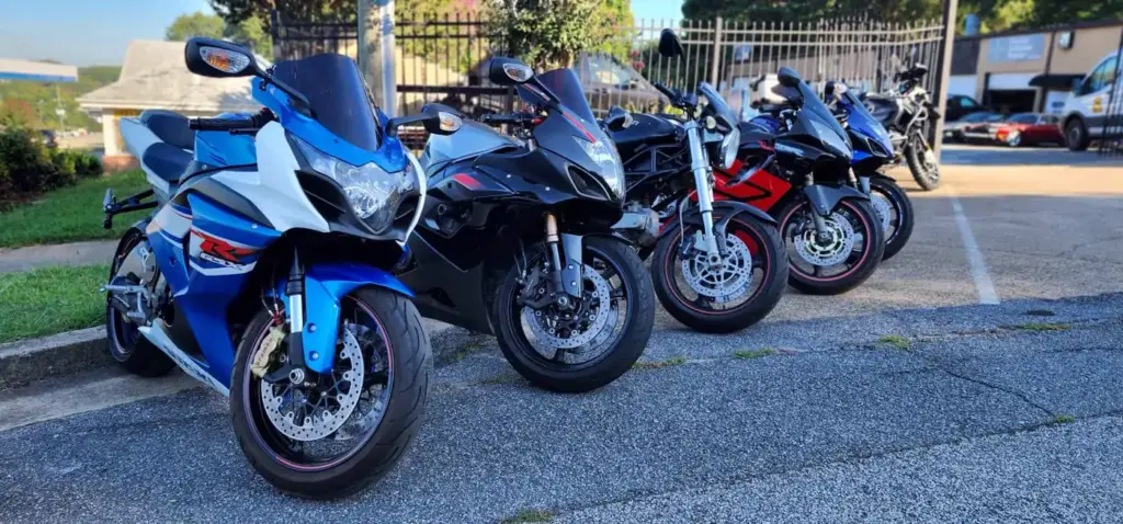 Four sport motorcycles are parked in a row on a paved lot by a building and black metal fence.