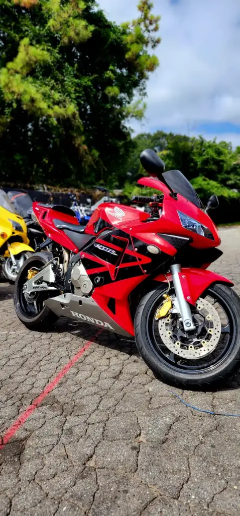 A red Honda motorcycle is parked on a cobblestone lot with trees and other motorcycles in the background.