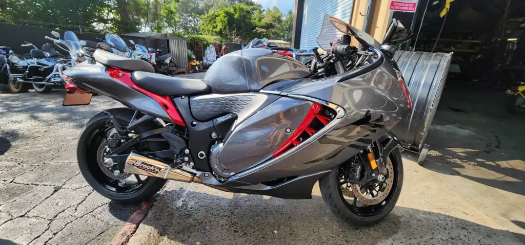 A sleek grey and red sport motorcycle is parked outdoors with other bikes and trees in the background.