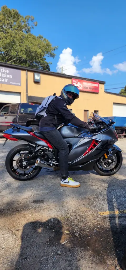 A person with a helmet and backpack sits on a gray sport motorcycle in a sunny parking lot.