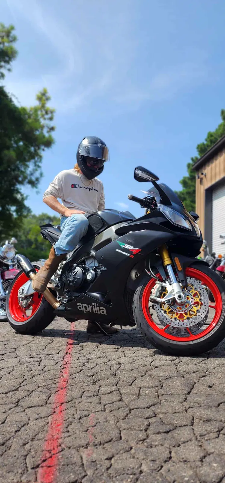 Independent motorcycle shop Acworth — helmeted rider on a black Aprilia RSV4 with red wheels in the shop lot
