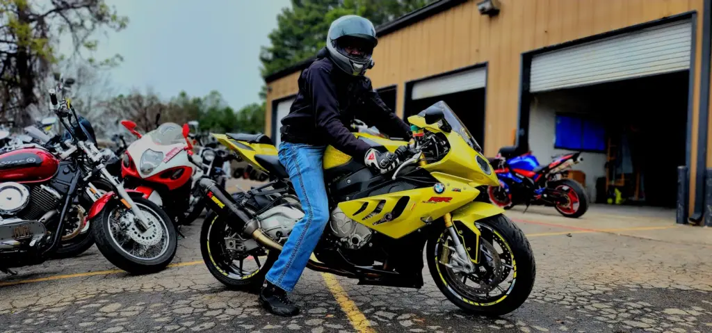 A person wearing a helmet and jeans sits on a yellow sport motorcycle in a parking lot by a garage.