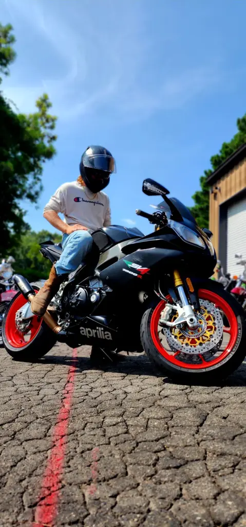 A person in a helmet and Champion shirt sits on a black Aprilia motorcycle with red wheels outdoors.
