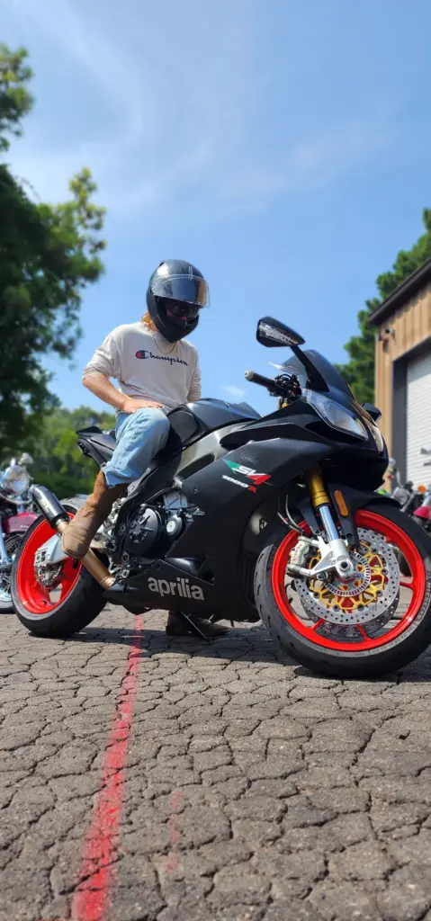 A person in a helmet rides a black Aprilia motorcycle with red rims outdoors on a sunny day.