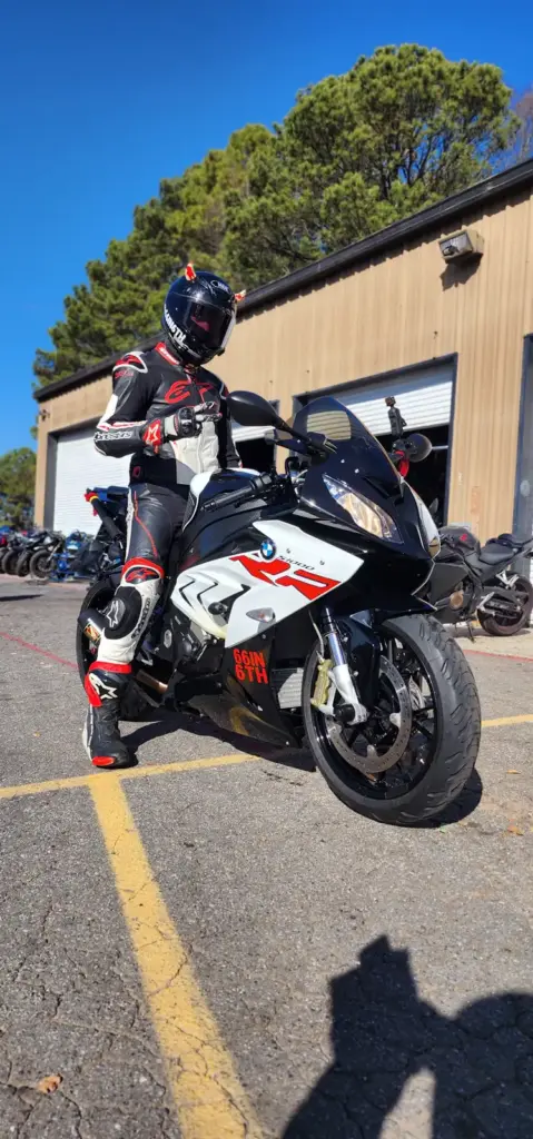 A motorcyclist in full gear sits on a black and white sport bike outside open garage buildings.