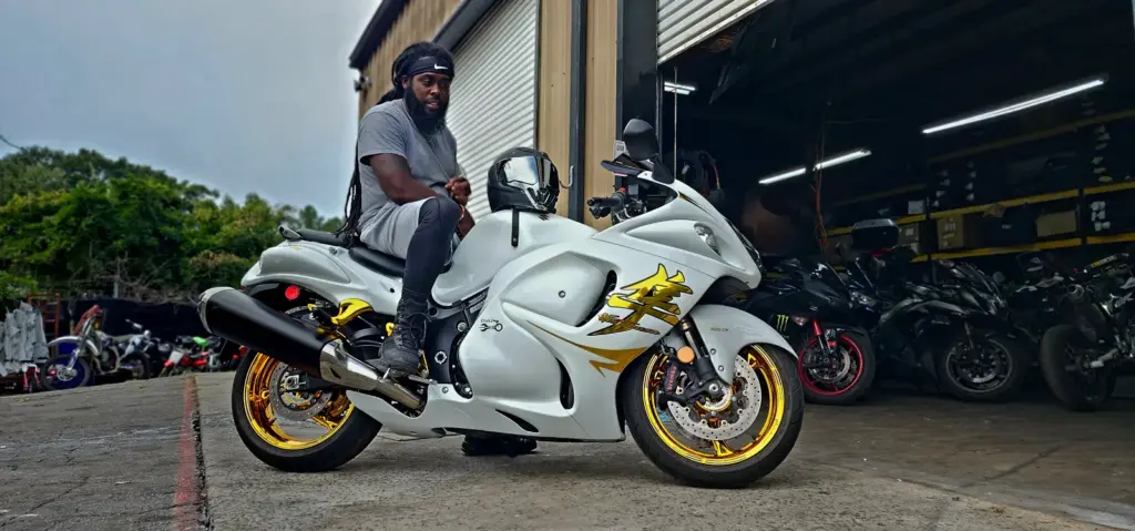 A man in gray athletic wear is seated on a white sport motorcycle with gold accents by a garage door.