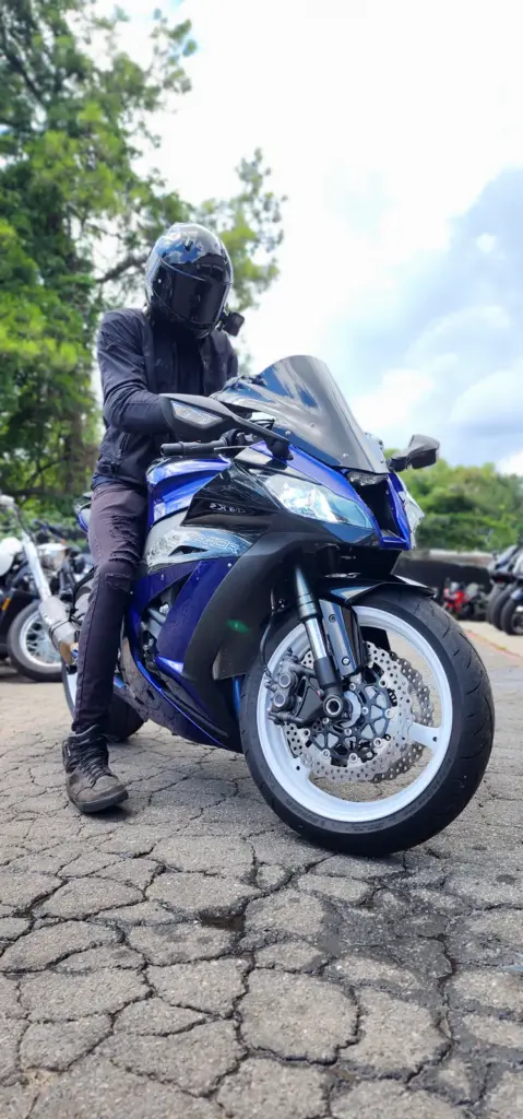 A person in black gear and helmet sits on a blue sport motorcycle in a parking area with trees nearby.