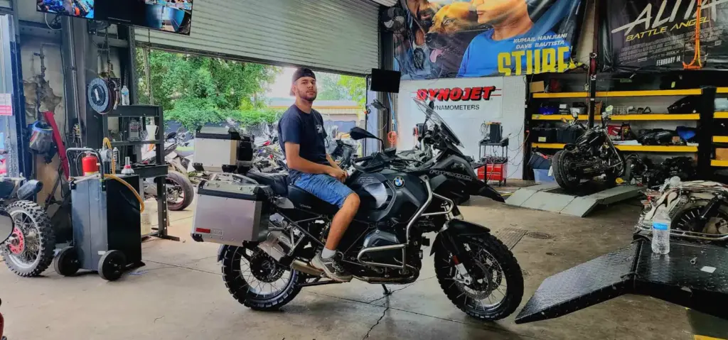 A man in a cap and shorts sits on a BMW adventure motorcycle in a busy garage workshop.
