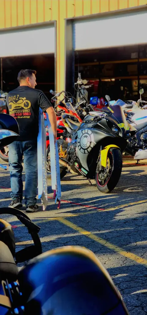 A man holds two motorcycle ramps outside a sunlit motorcycle shop near parked bikes in the lot.
