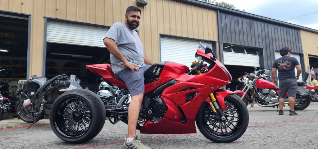 A man wearing gray sits on a red three-wheeled motorcycle outside a garage with several bikes nearby.