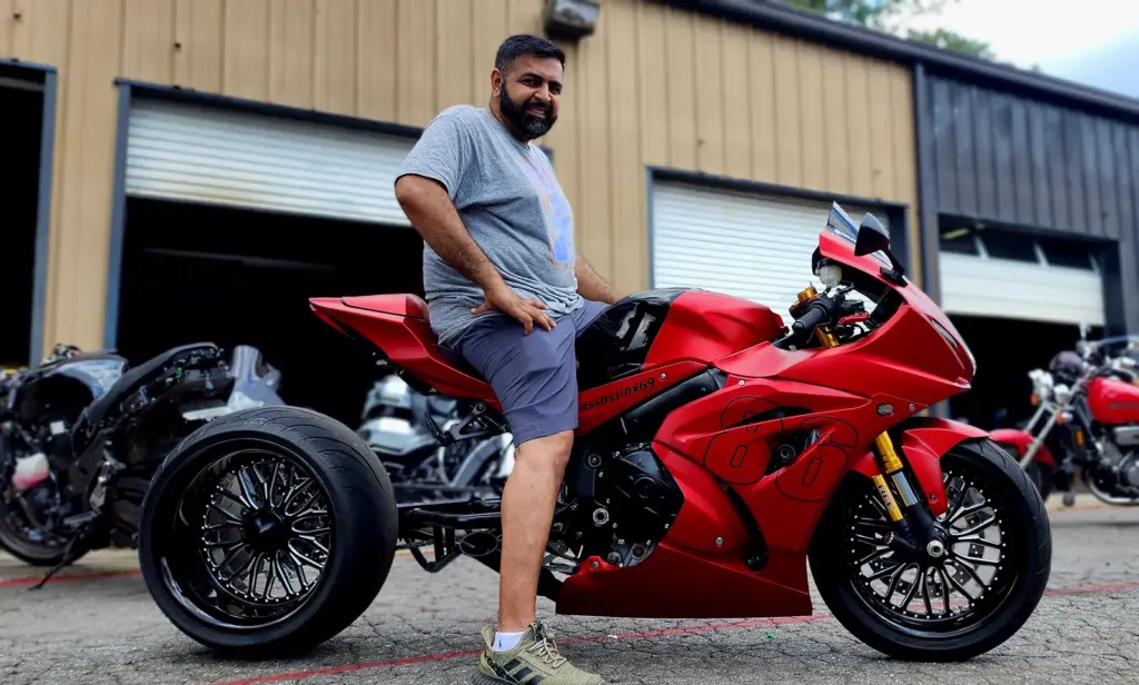 A bearded man in a gray shirt sits on a red sport motorcycle outside an industrial garage.