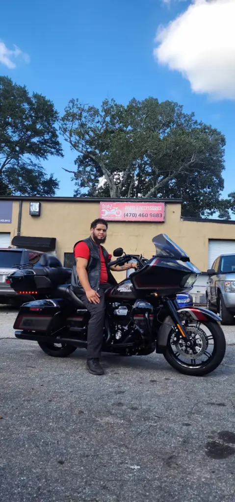 A man wearing a red shirt sits on a black motorcycle in a parking lot near an auto accessories shop.
