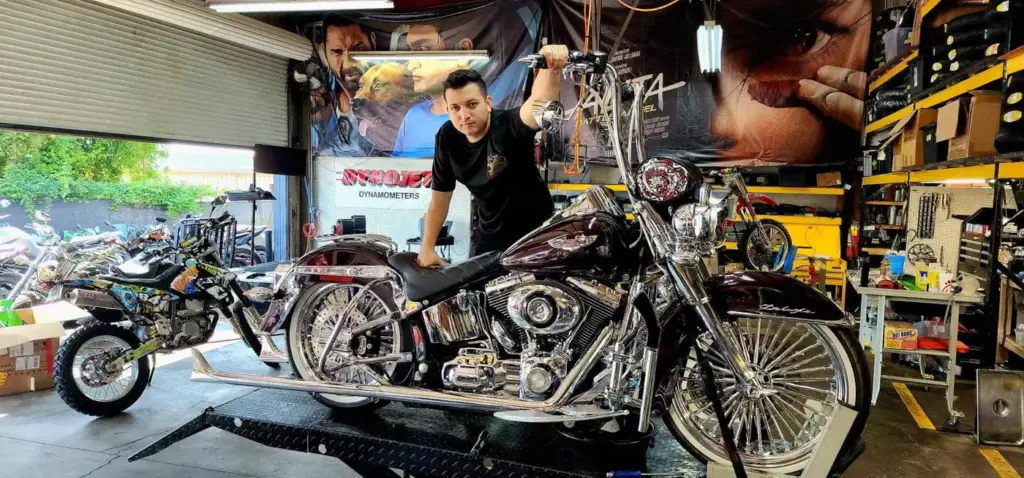 A man poses with a shiny custom motorcycle in a workshop filled with tools and other bikes.