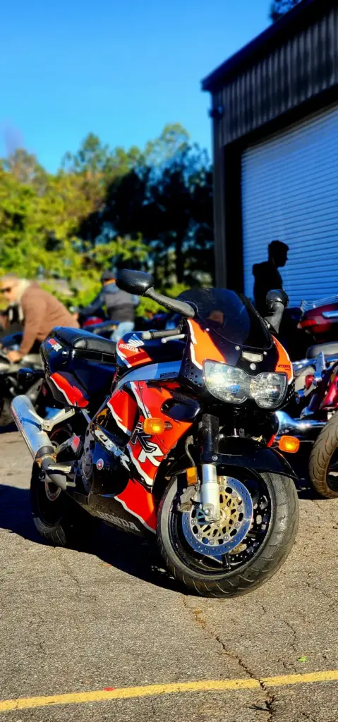 A red and black sport motorcycle is parked in sunlight among other bikes, with people visible behind.