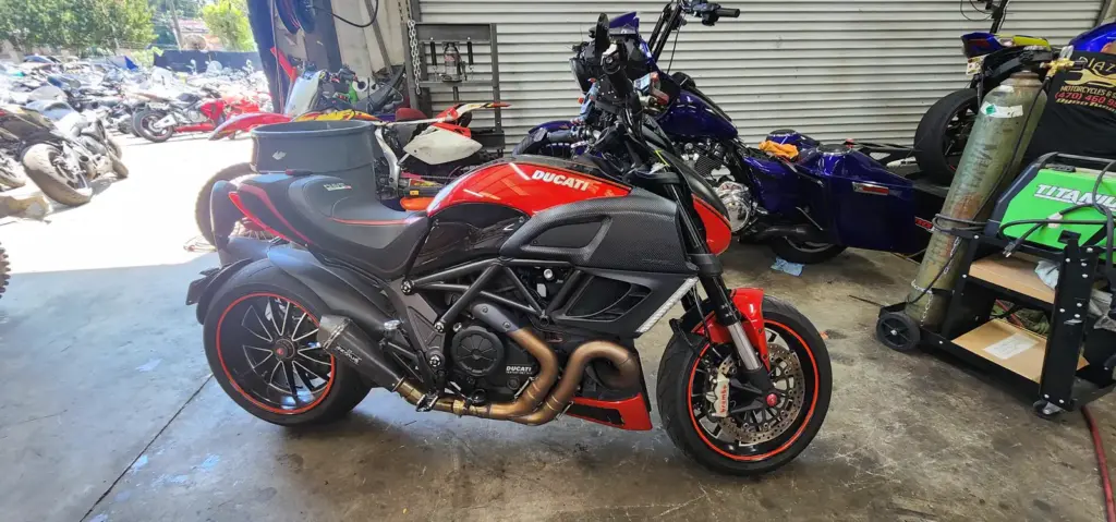 A red and black Ducati motorcycle is parked in a garage with other bikes and assorted tools visible.
