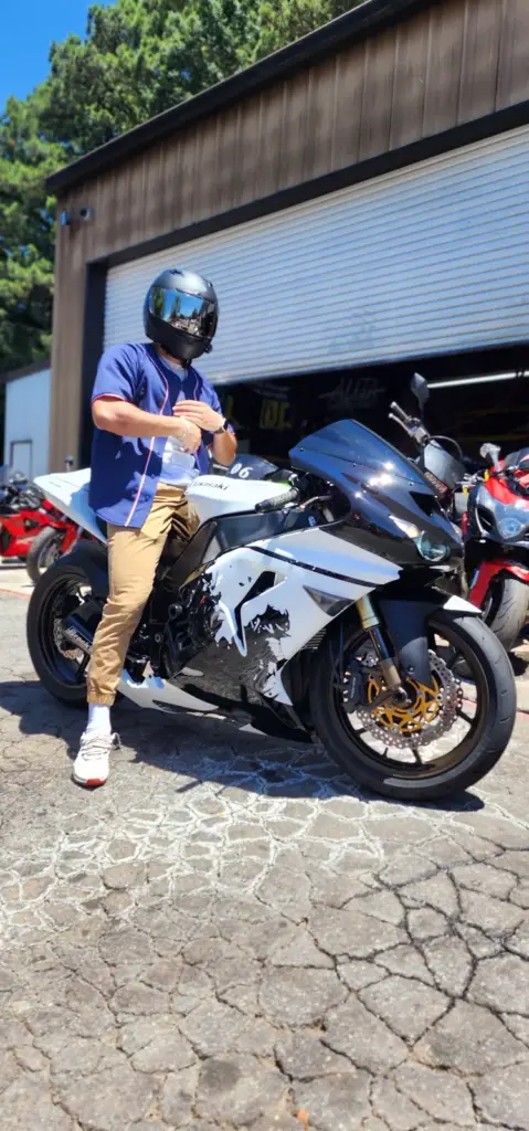 A person in a helmet and blue shirt sits on a black and white motorcycle outside a garage with bikes.
