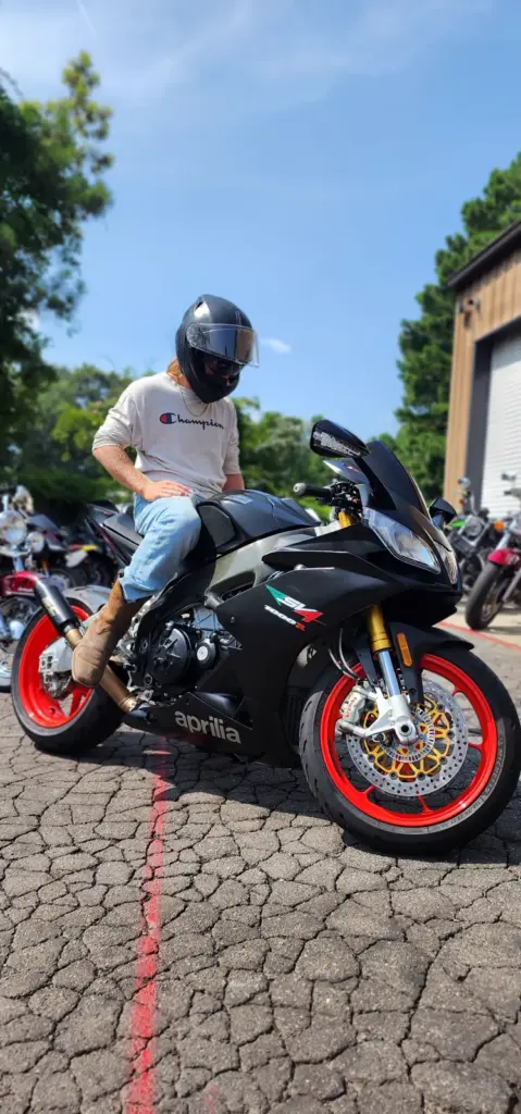A person in a helmet and boots sits on a black Aprilia motorcycle with red rims in a parking lot.