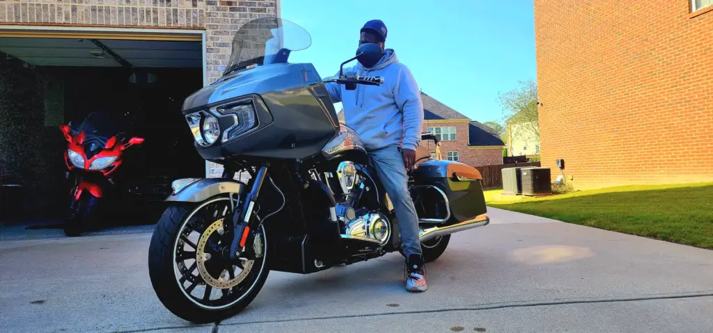 A person in a hoodie and cap stands by a large motorcycle in a suburban driveway under sunny skies.