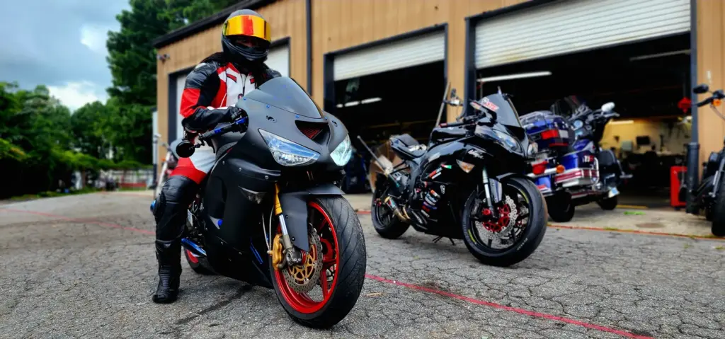 Rider in helmet and gear sits on a black sport motorcycle near a garage, with bikes visible behind.
