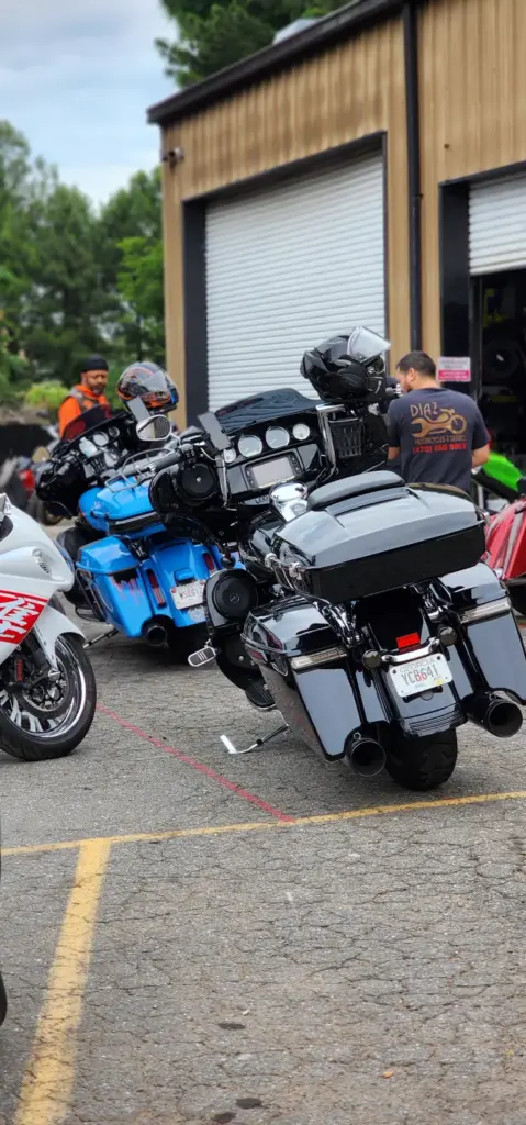 Motorcycles are parked outside a garage while people in helmets and riding gear stand nearby.