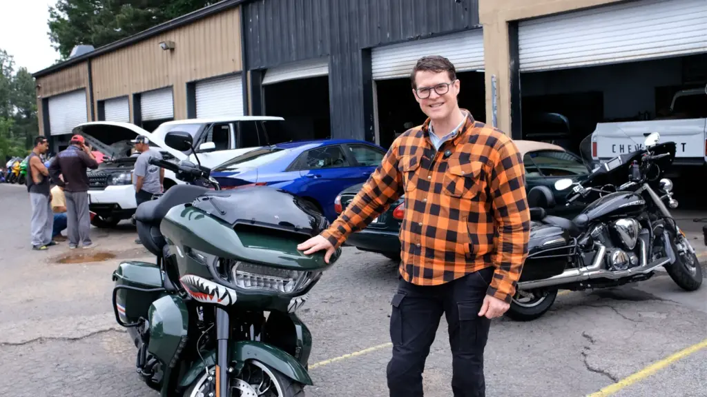 A smiling man in a plaid shirt poses by a green motorcycle with shark mouth art in a parking lot.