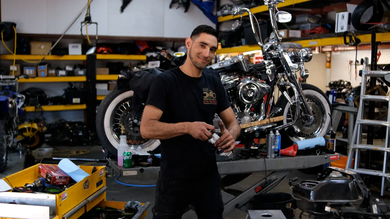 Motorcycle transport in Woodstock — smiling Diaz technician holding a part beside a chrome Harley softail on a lift