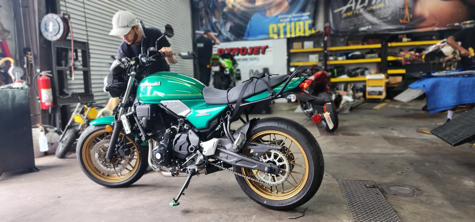 Stranded motorcycle pickup Woodstock — teal-green retro Kawasaki with gold spoked wheels on the Diaz shop floor being worked on