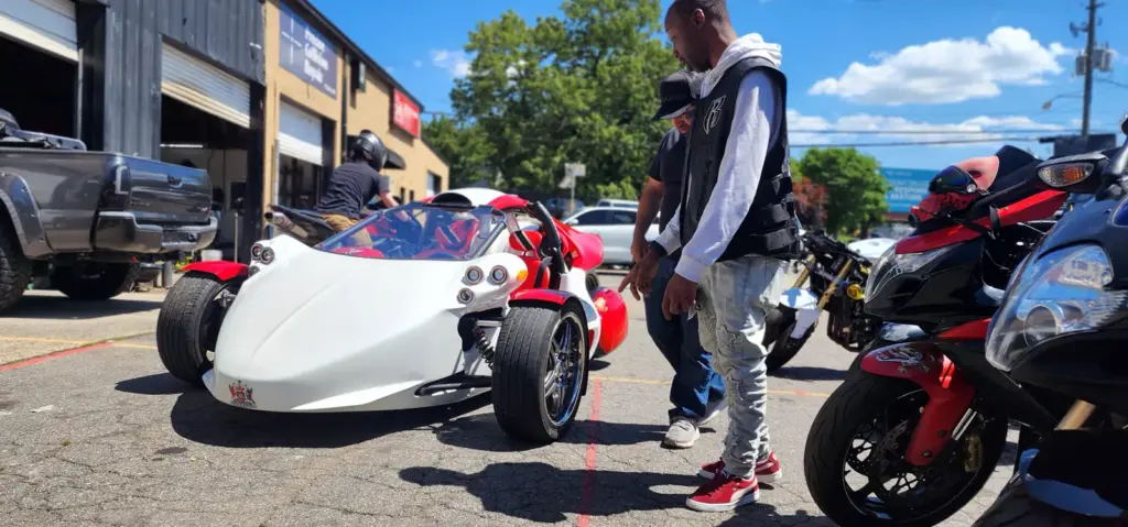 A man stands by a white three-wheeled car in a parking lot, surrounded by motorcycles and people.