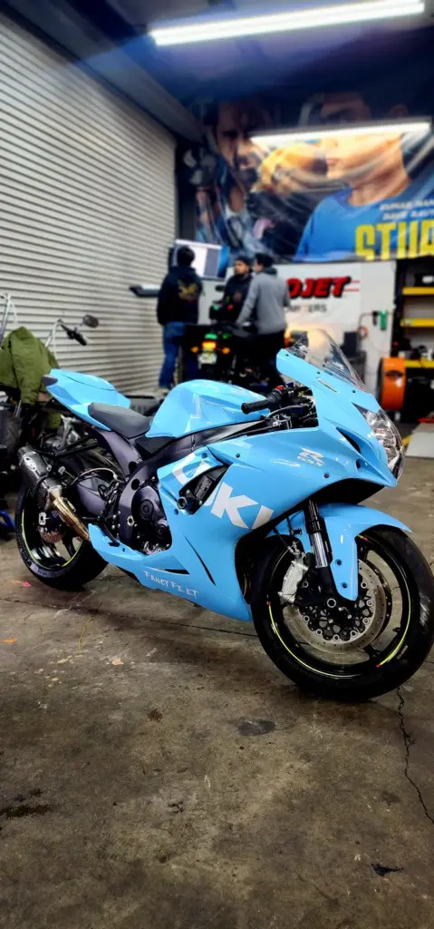 A shiny blue sport motorcycle is parked in a garage, with people behind it and a mural on the wall.