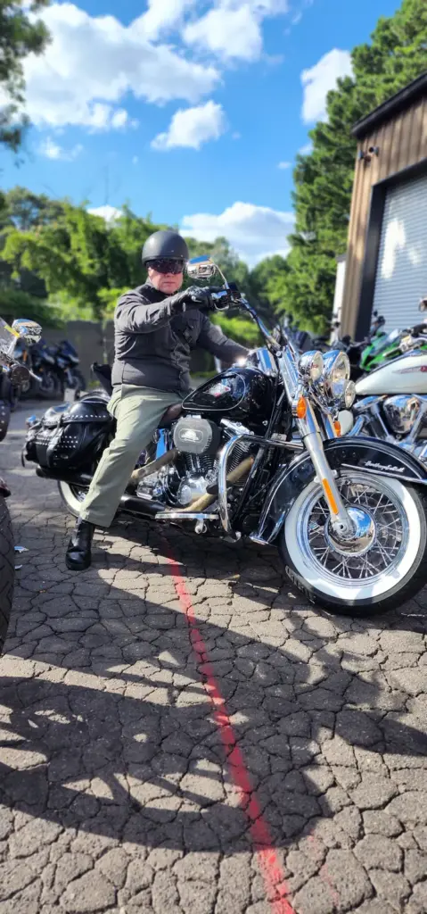 A person with a helmet and sunglasses sits on a black motorcycle in a parking lot on a sunny day.