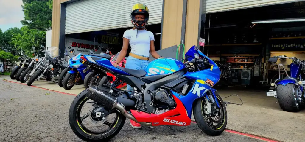 A person in a helmet stands behind a blue Suzuki sportbike near a motorcycle garage with other bikes.