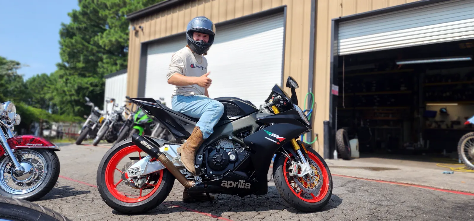 motorcycle transport cost Canton — young rider in a helmet giving a thumbs-up on a black Aprilia RSV4 outside the shop bays