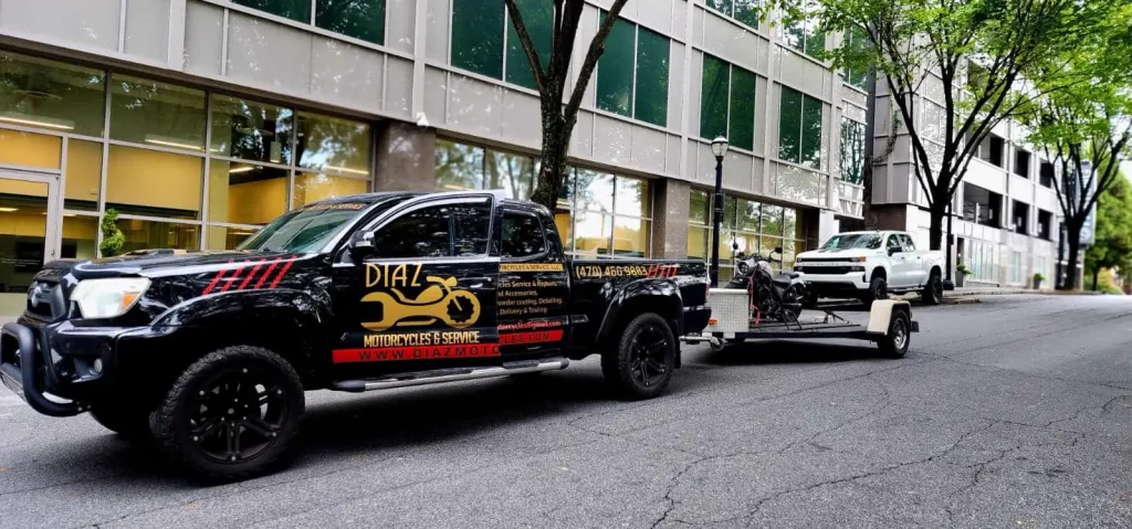 The black branded Diaz pickup with an open trailer carrying a bike parked on a city street beside an office building.