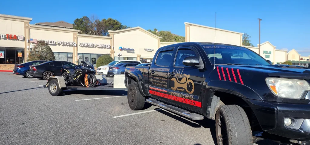Black Diaz-branded Toyota Tacoma towing an open trailer with a black sportbike strapped down, parked in a strip mall lot.