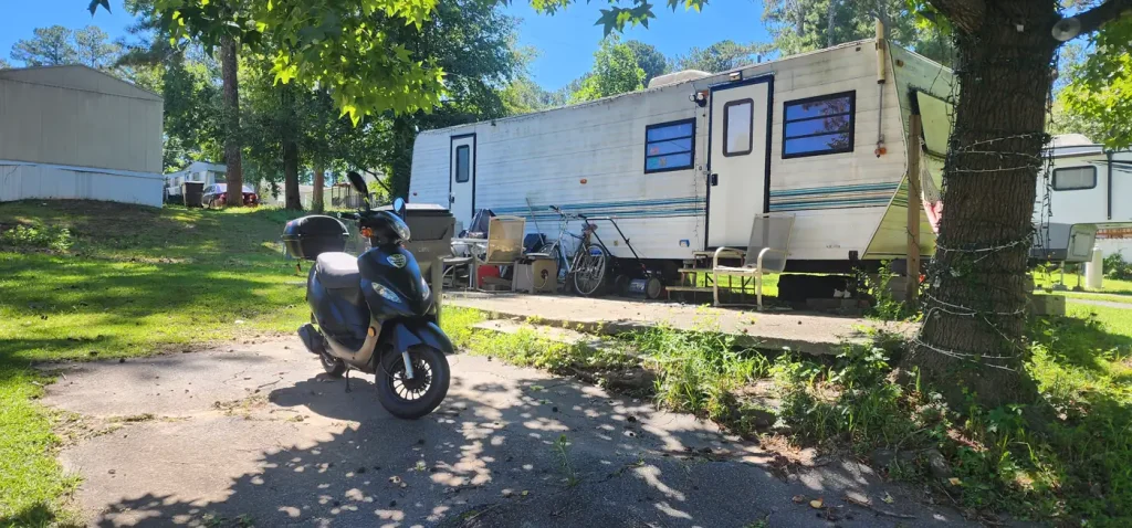 Black scooter parked on a concrete pad outside a white travel trailer in a wooded residential lot on a sunny day.