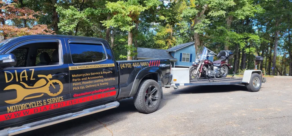 Black Toyota Tacoma with full Diaz Motorcycles door graphics towing an open flatbed trailer carrying a maroon Harley-Davidson cruiser in a wooded residential driveway.