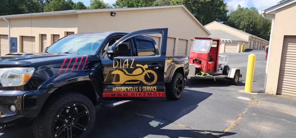 A black pickup truck with a “Diaz Motorcycles & Service” logo tows a small red vehicle on a trailer.