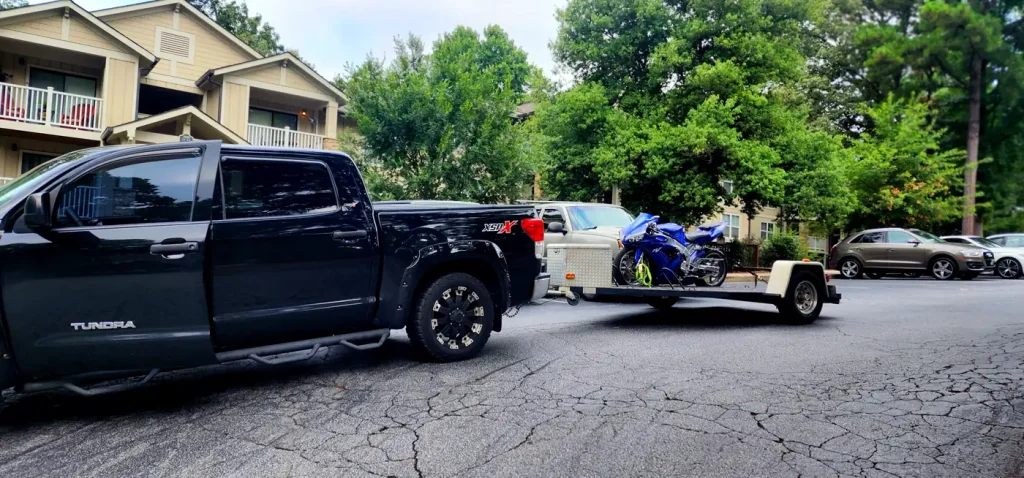 Black Toyota Tundra pickup towing an open single-rail trailer with a blue Yamaha R6 strapped on, parked in an apartment complex lot.