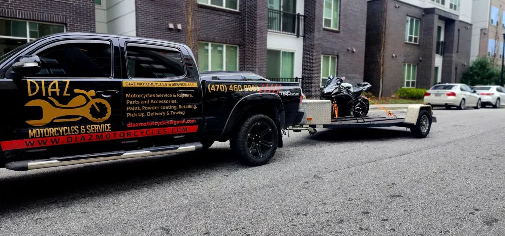 Black wrapped Diaz Tacoma with open trailer carrying a black sportbike, parked curbside at a modern apartment building.