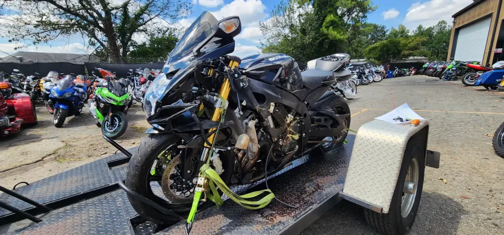 Damaged black Suzuki GSX-R with stripped fairings and exposed wiring strapped to an open trailer with green ratchet straps in the shop lot.