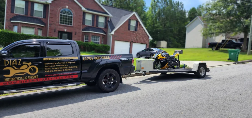 Diaz-branded black Tacoma towing an open trailer with a yellow and black sportbike strapped down, parked on a suburban street.