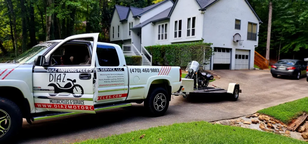 Diaz Motorcycles branded white GMC pickup with an open single-rail trailer carrying a cruiser, parked in a residential driveway in front of a white house.