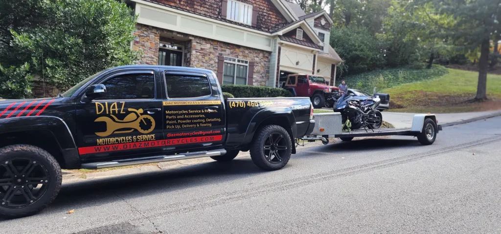 Diaz-wrapped black Toyota Tacoma towing an open trailer with a black sportbike loaded, parked outside a suburban brick home.
