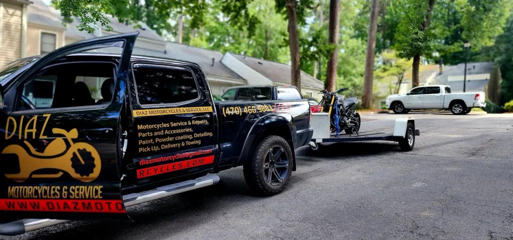 Diaz-wrapped Tacoma with door open towing a blue supermoto/dirt bike on an open trailer in a wooded neighborhood