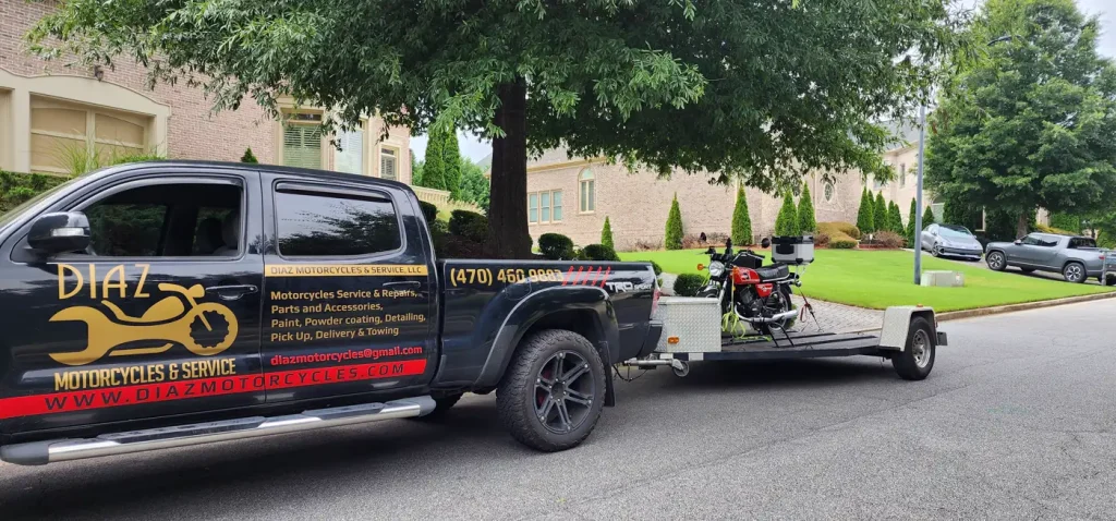 Diaz-wrapped Tacoma with an open trailer carrying a red classic standard motorcycle, parked in a brick-home driveway.