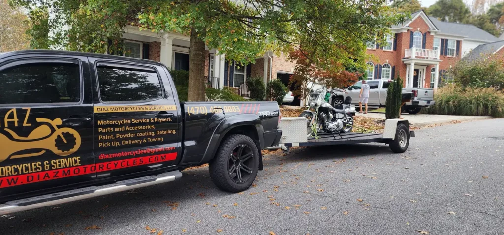 Diaz-wrapped Tacoma towing an open trailer with a chrome Harley cruiser loaded, parked on a suburban street with fall leaves.
