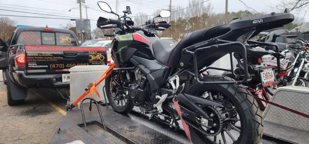 A black motorcycle is strapped to a trailer beside a service truck in a parking lot, ready for transport.