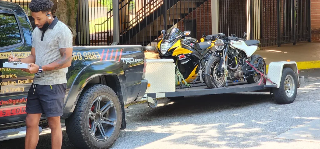Man with a clipboard writing notes beside the Diaz Tacoma and loaded trailer carrying a yellow GSX-R and a black cafe-style bike.